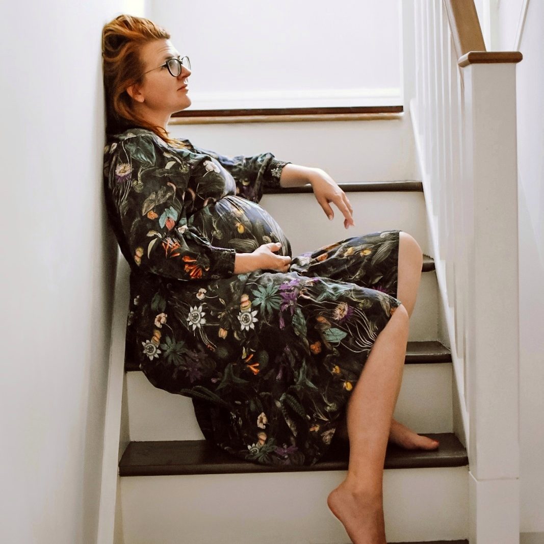 A pregnant woman sitting on a staircase, wearing a floral dress and glasses, with a relaxed expression.