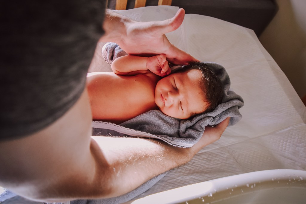 A close-up of a person gently holding a newborn baby wrapped in a gray towel, captured in soft natural light.