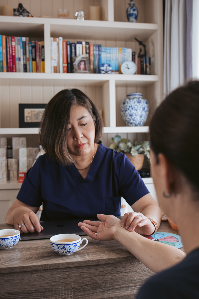 A woman in a navy uniform examines the wrist of another person in a cozy indoor setting, with shelves of books and decorative items in the background.