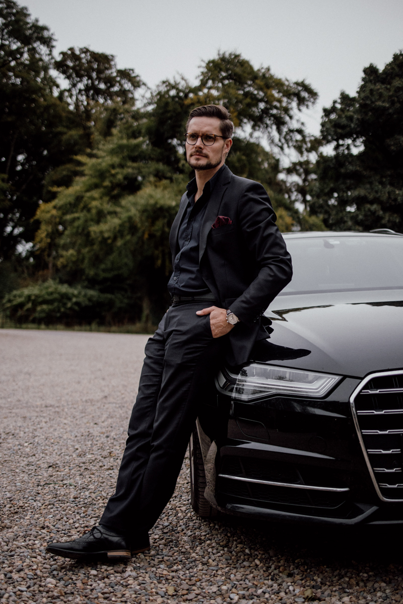 A man in a black suit and glasses leaning against a luxury car in an outdoor setting, surrounded by greenery.