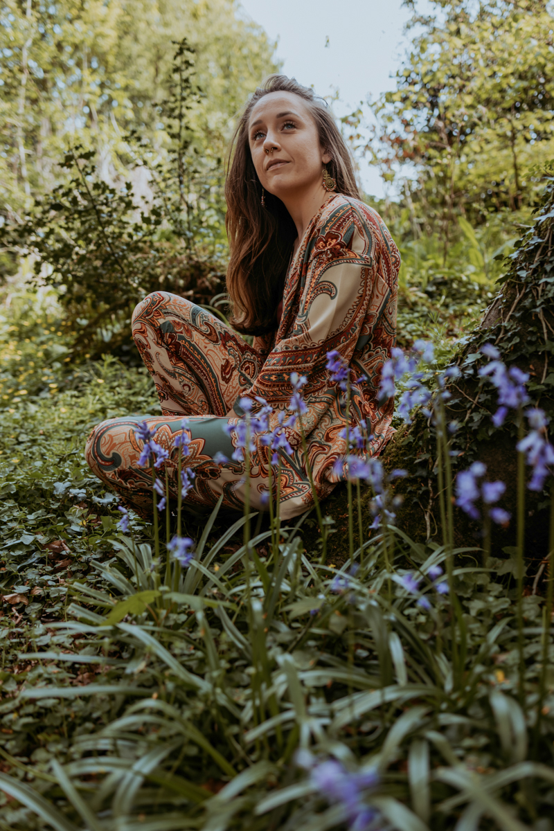 A woman sitting in a lush green environment surrounded by flowers, wearing a colorful patterned outfit, gazing thoughtfully.
