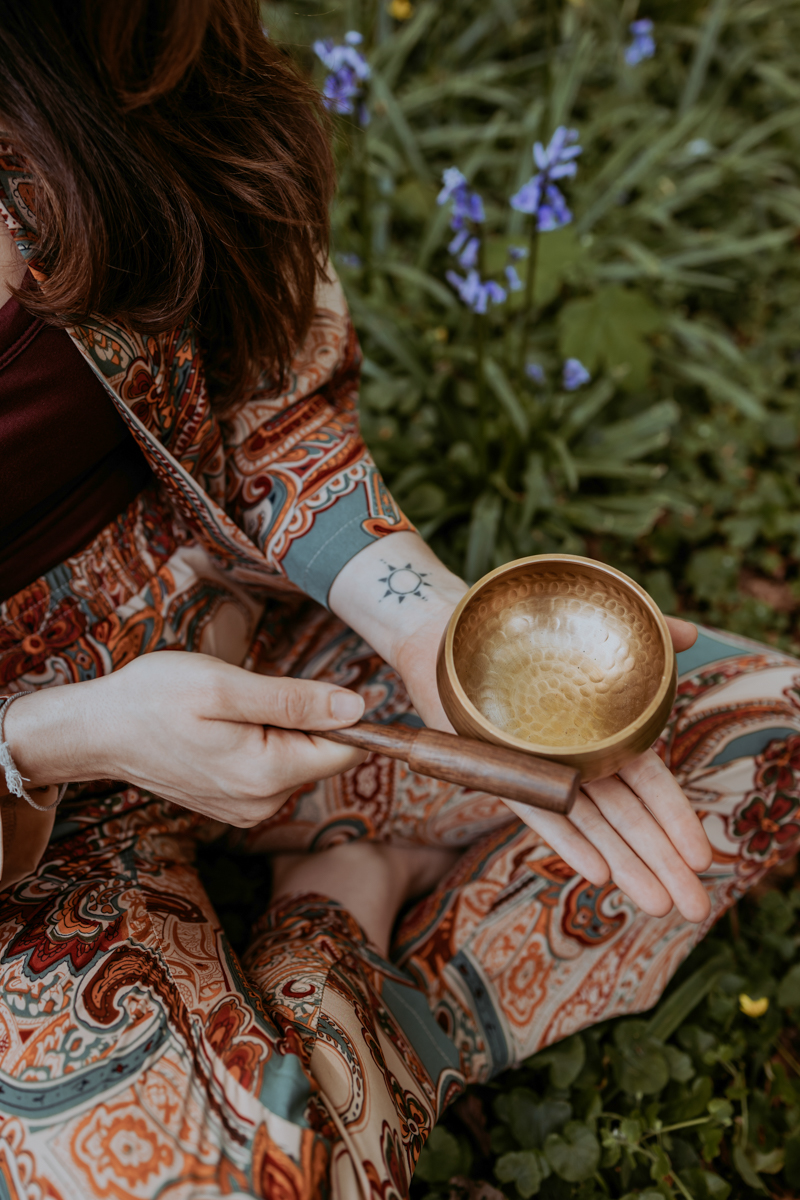 A person seated amidst greenery holds a brass singing bowl and mallet, wearing a colorful patterned outfit, reflecting a serene lifestyle.