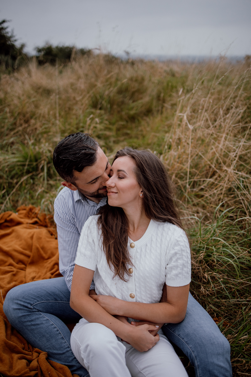 A couple sitting on a blanket in a grassy field, sharing an intimate moment, with the man leaning in to kiss the woman while she smiles softly.