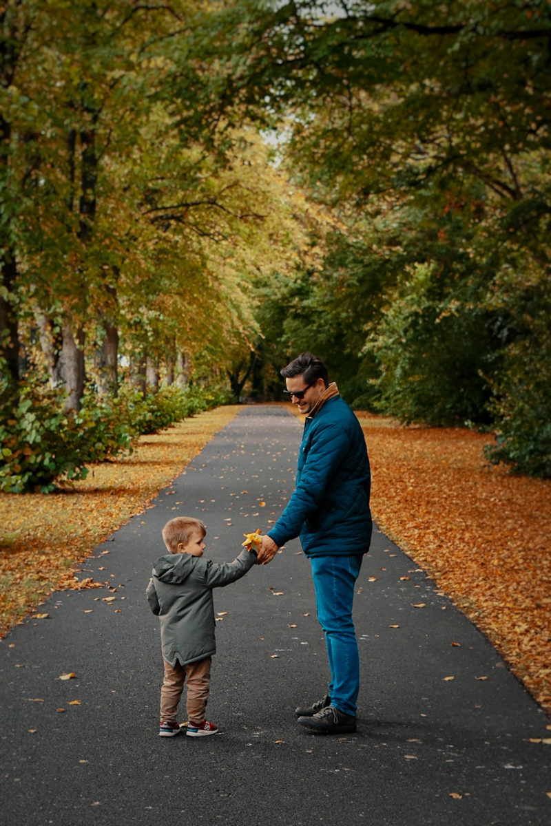 A father and son holding hands while walking down a tree-lined path covered with autumn leaves, exchanging a moment of joy.
