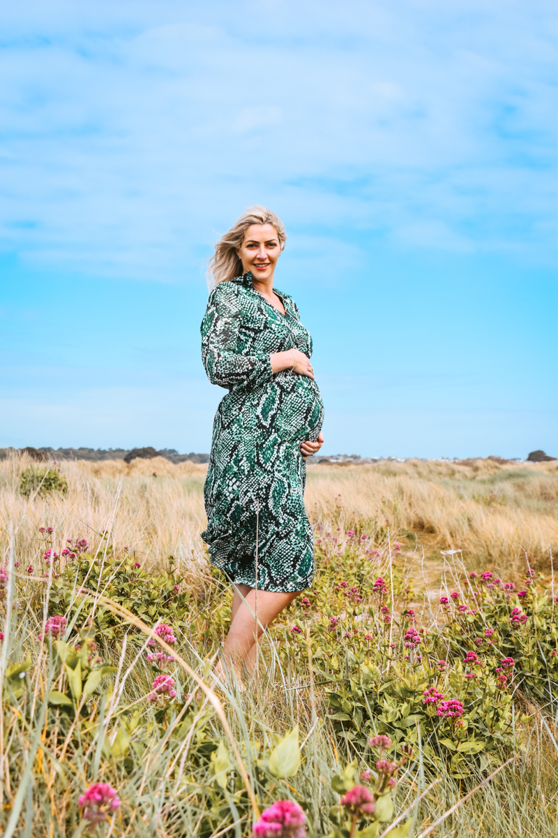 Pregnant woman standing in a field of tall grass and pink flowers, wearing a green patterned dress, against a blue sky.