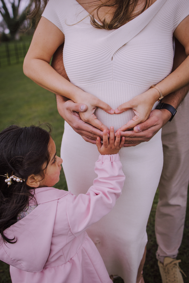 A maternity photo featuring a woman in a light-colored dress, holding her belly with her partner's hands forming a heart shape around it, while a child reaches out to touch the belly.