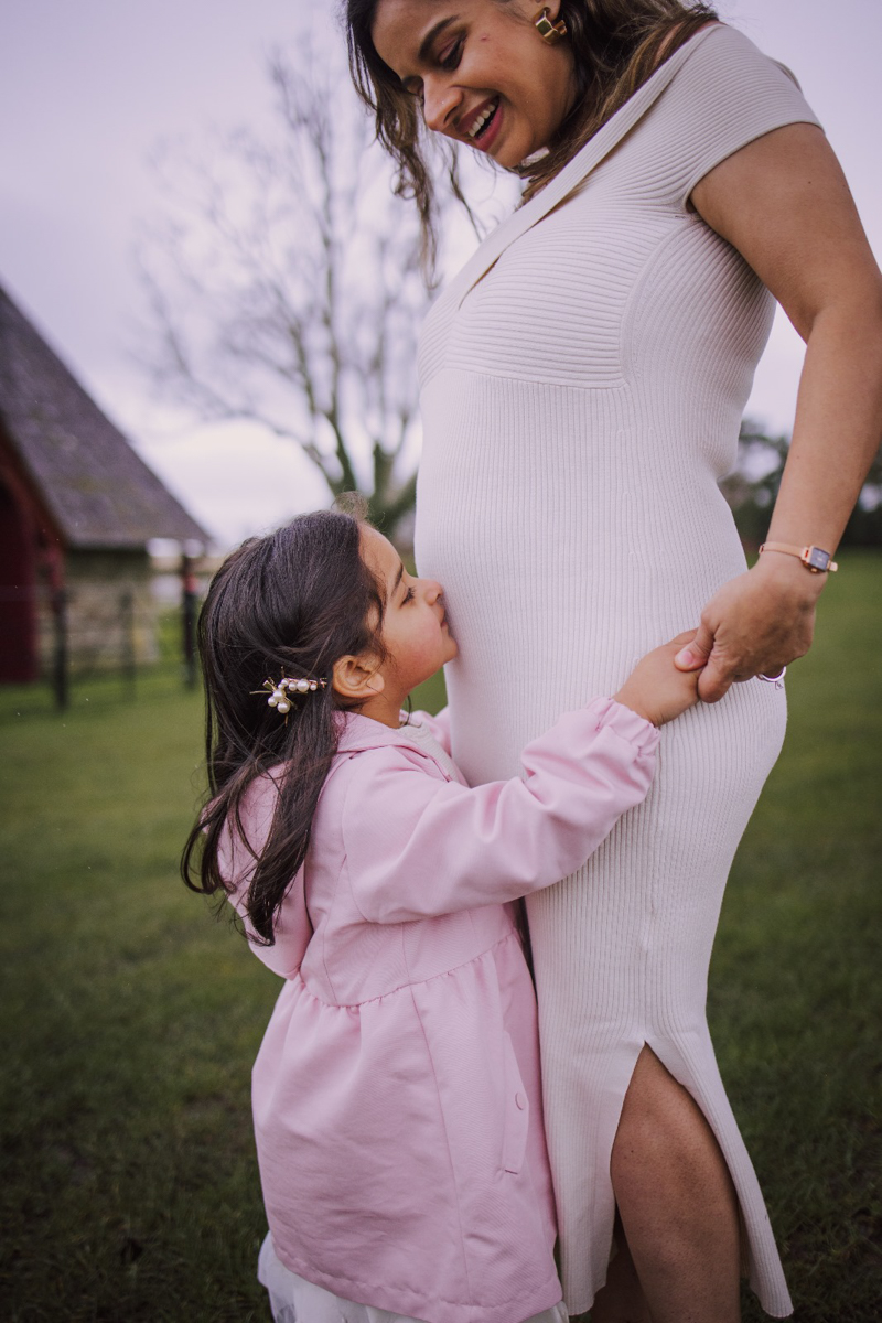 A pregnant woman in a white dress smiles down at a young girl in a pink coat, who is playfully holding her belly in a picturesque outdoor setting.
