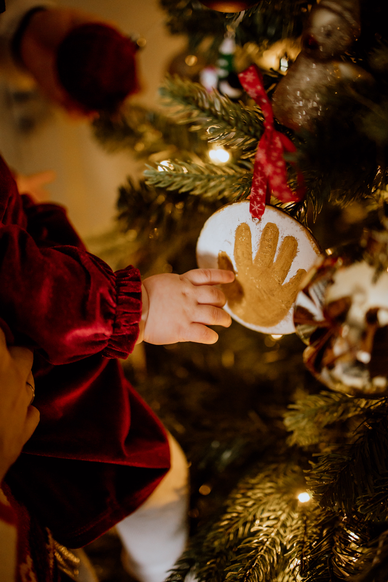 A close-up of a child's hand reaching towards a decorated Christmas tree ornament, featuring a handprint design, with warm lighting creating a cozy atmosphere.