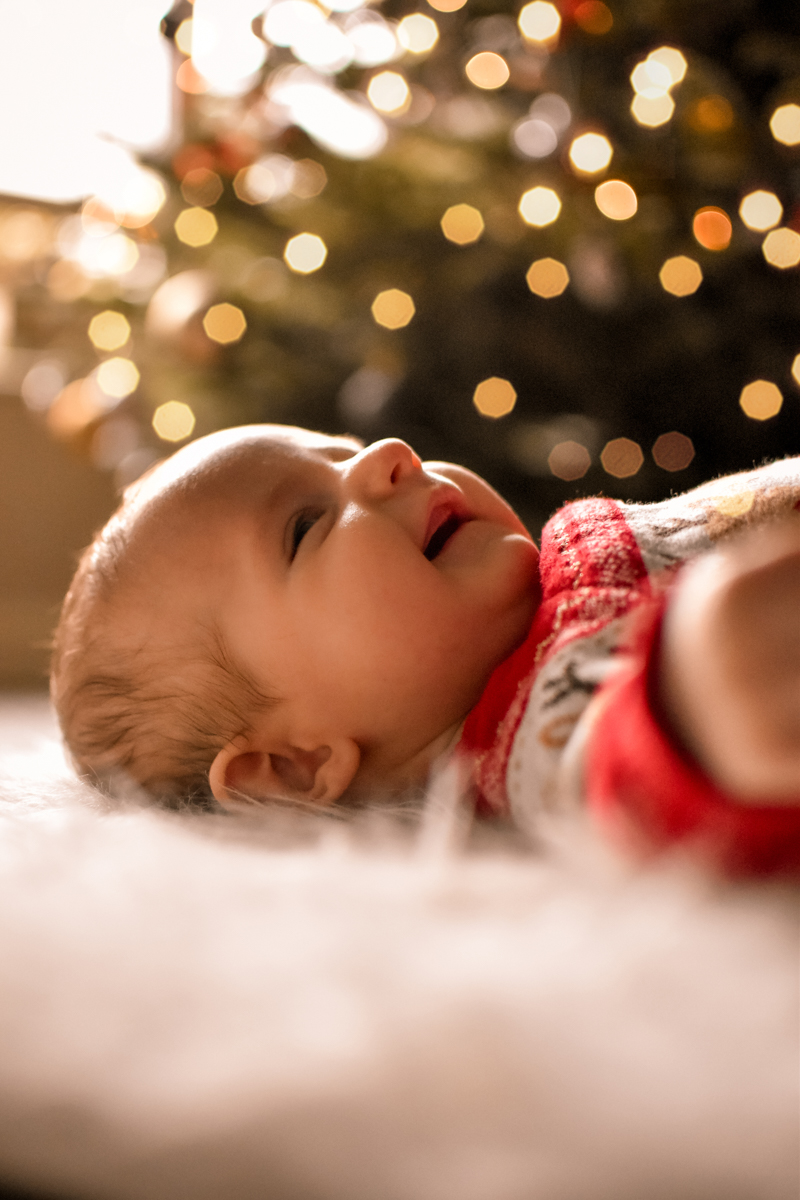A smiling baby lying on a soft surface with Christmas lights blurred in the background.