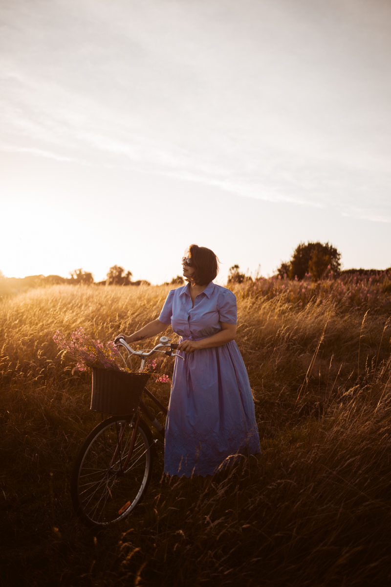 A woman in a blue dress standing beside a bicycle with flowers in the basket, set against a golden sunset in a field.