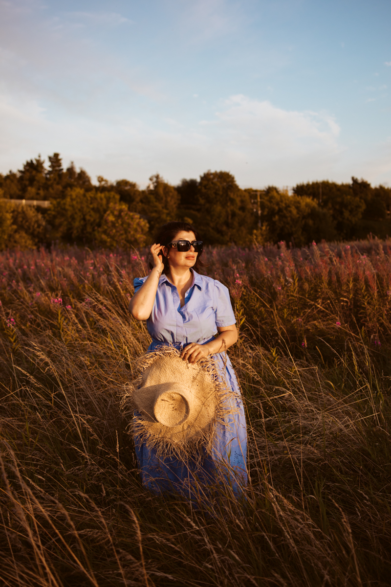 A woman in a blue dress stands in a field of tall grass and wildflowers, holding a straw hat and gazing thoughtfully into the distance as the sun sets behind her.