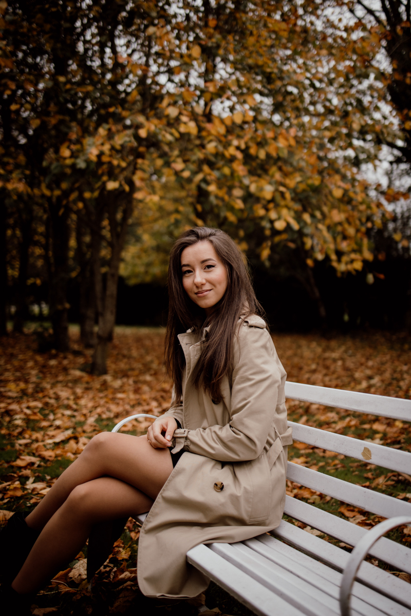 Woman sitting on a park bench surrounded by autumn leaves, wearing a beige trench coat, exuding confidence and tranquility.