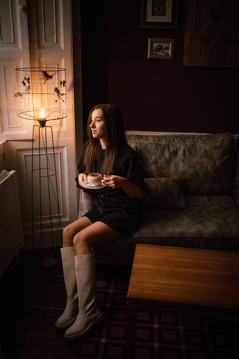 A woman sitting on a couch holding a cup of coffee, wearing a short dress and knee-high boots, with a lamp and dark wall in the background.