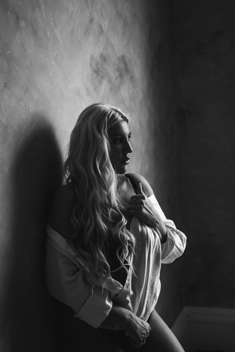 Black and white photo of a woman with long, flowing hair, wearing an open shirt, leaning against a textured wall. She gazes thoughtfully in profile, exuding confidence and softness.