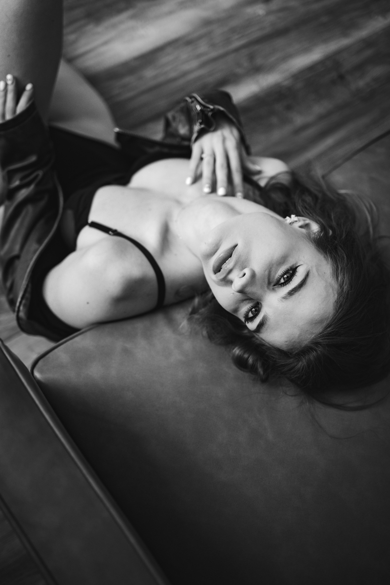 A woman lying on a leather surface, wearing a leather jacket and a black bra, gazing at the camera with soulful eyes in a black and white boudoir style photograph.