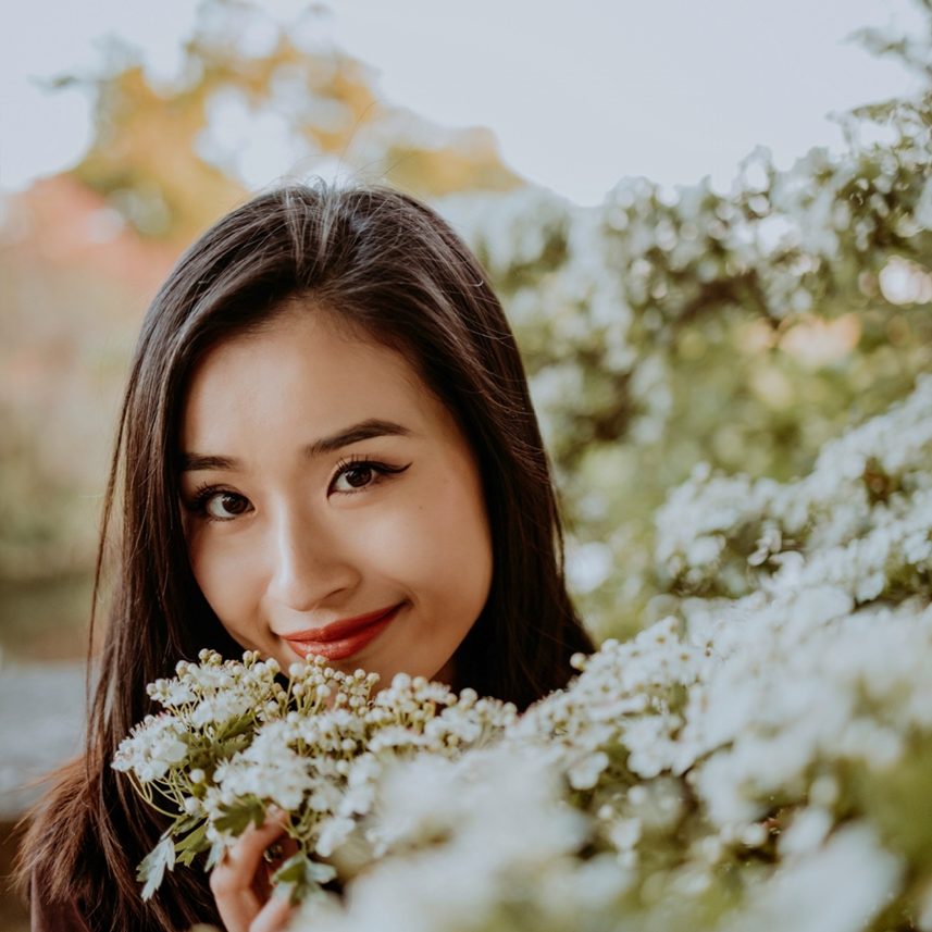 A smiling woman holding a bouquet of white flowers, surrounded by greenery, with a soft background.