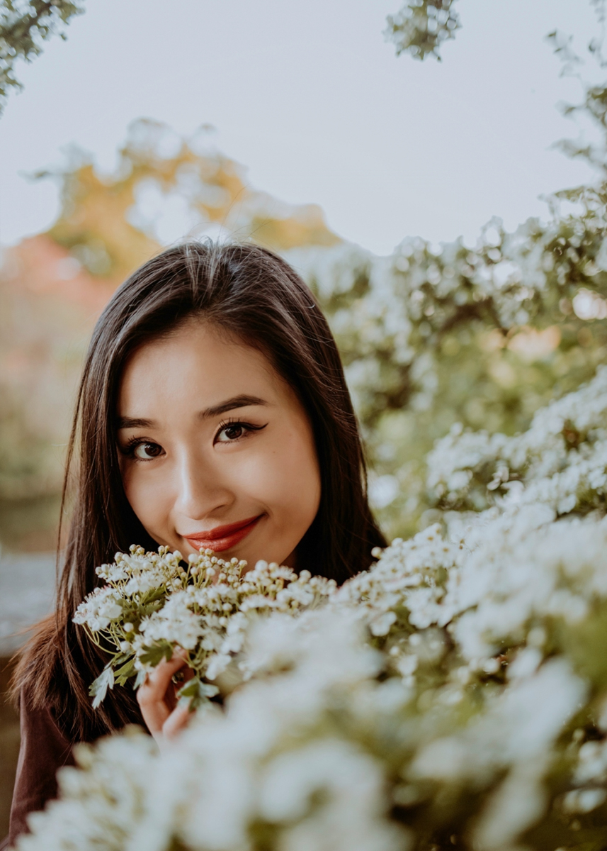 A woman smiling while holding white flowers, surrounded by greenery.