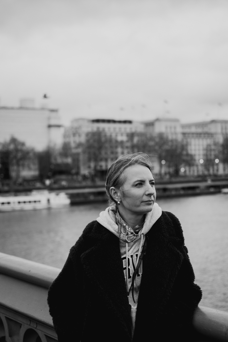A woman with short hair stands on a bridge overlooking a river, wearing a cozy coat and a hoodie, with a thoughtful expression against a cloudy sky.