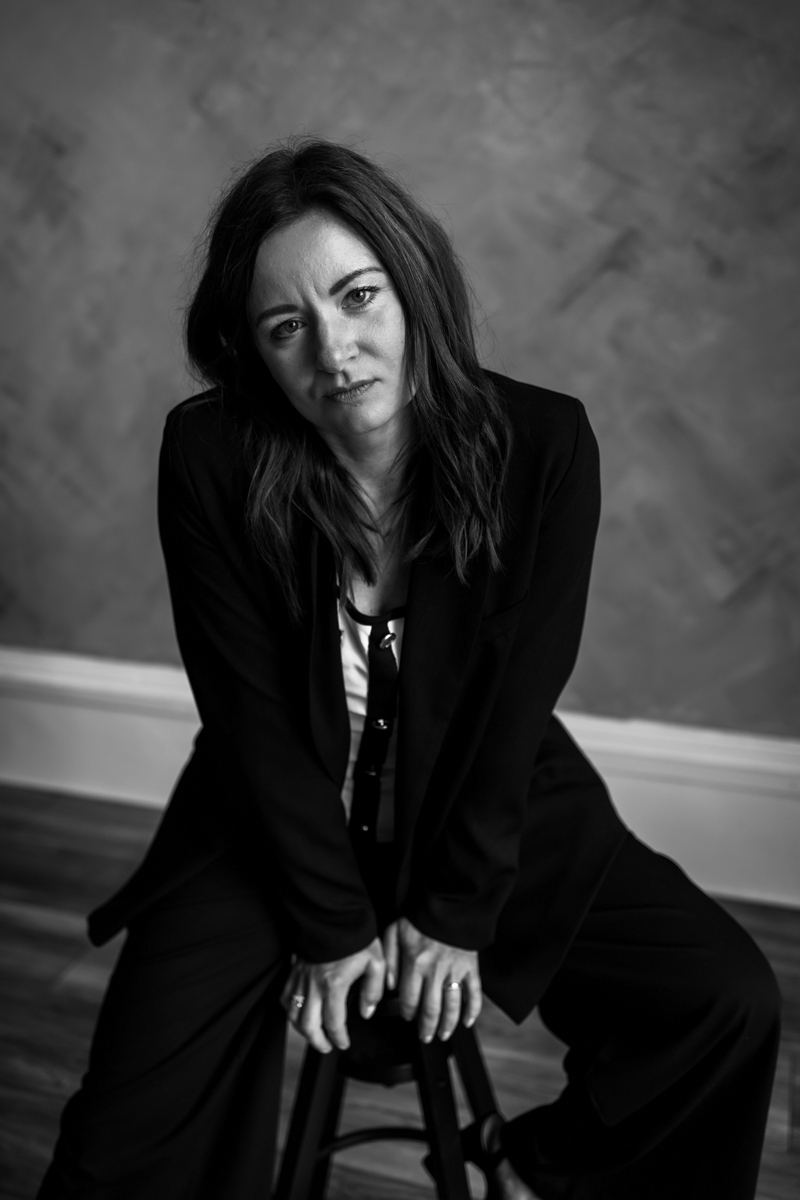 A woman in a black suit sitting on a stool, looking serious and thoughtful, with long hair and a neutral background.