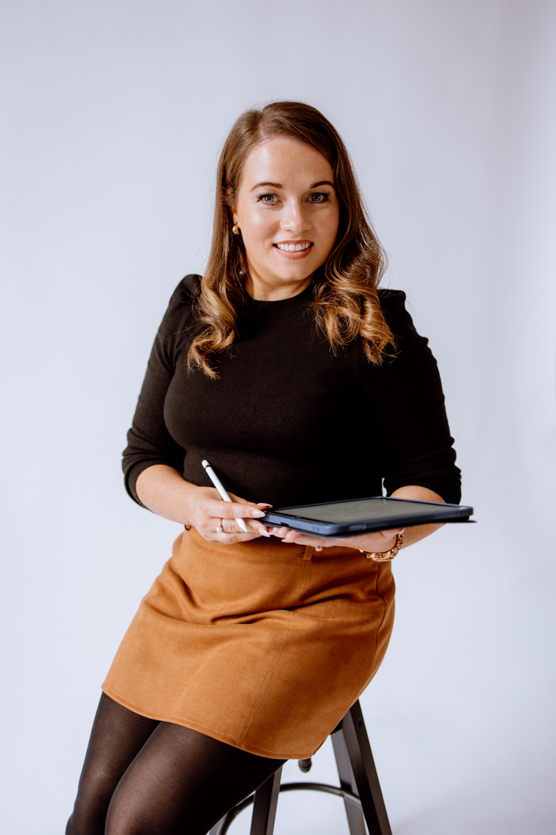 A woman sitting on a stool, holding a tablet and stylus, smiling at the camera, wearing a black top and a brown skirt against a plain white background.