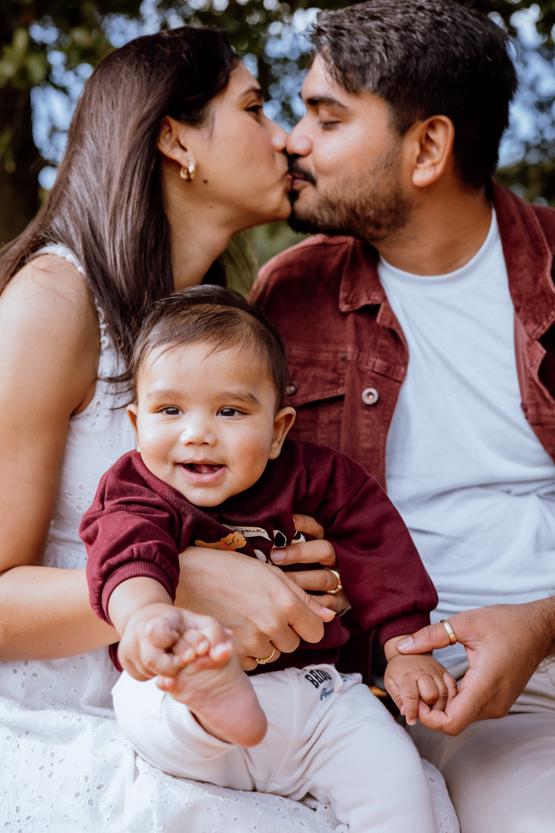 A happy baby smiling while being held by a woman and a man, who are kissing each other. The setting appears to be outdoors with greenery in the background.