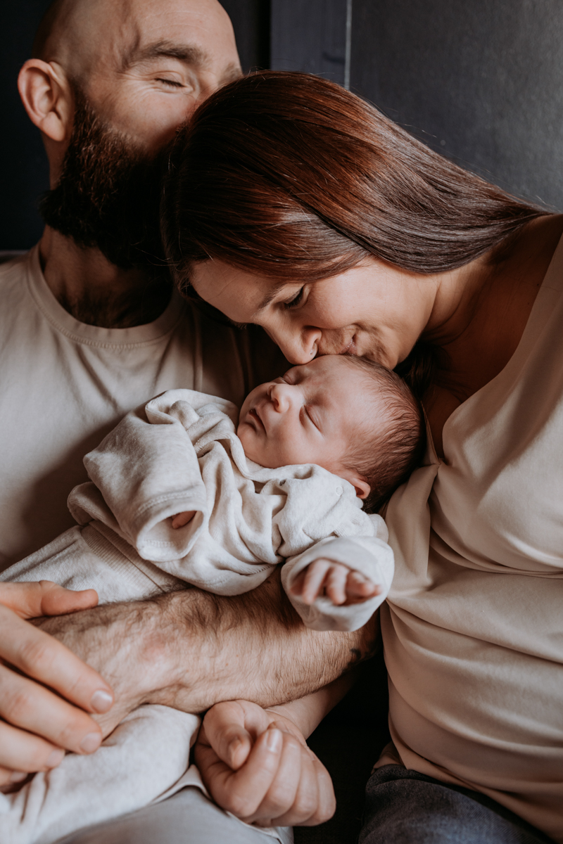 A loving couple holding a newborn baby, with the mother kissing the baby's forehead and the father leaning in closely. The family appears warm and affectionate, showcasing a tender moment.
