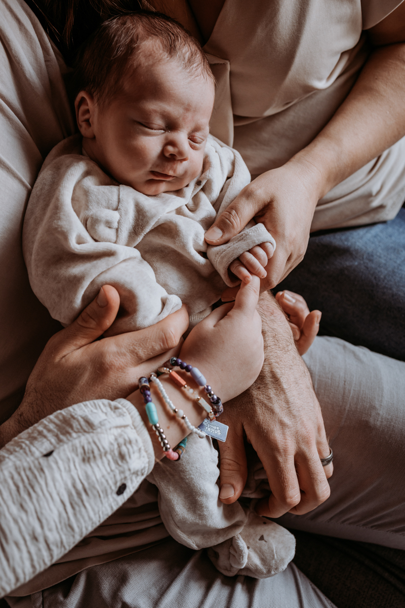 A close-up of a baby peacefully sleeping in the arms of adults, with hands gently holding the baby and colorful bracelets visible on one wrist.