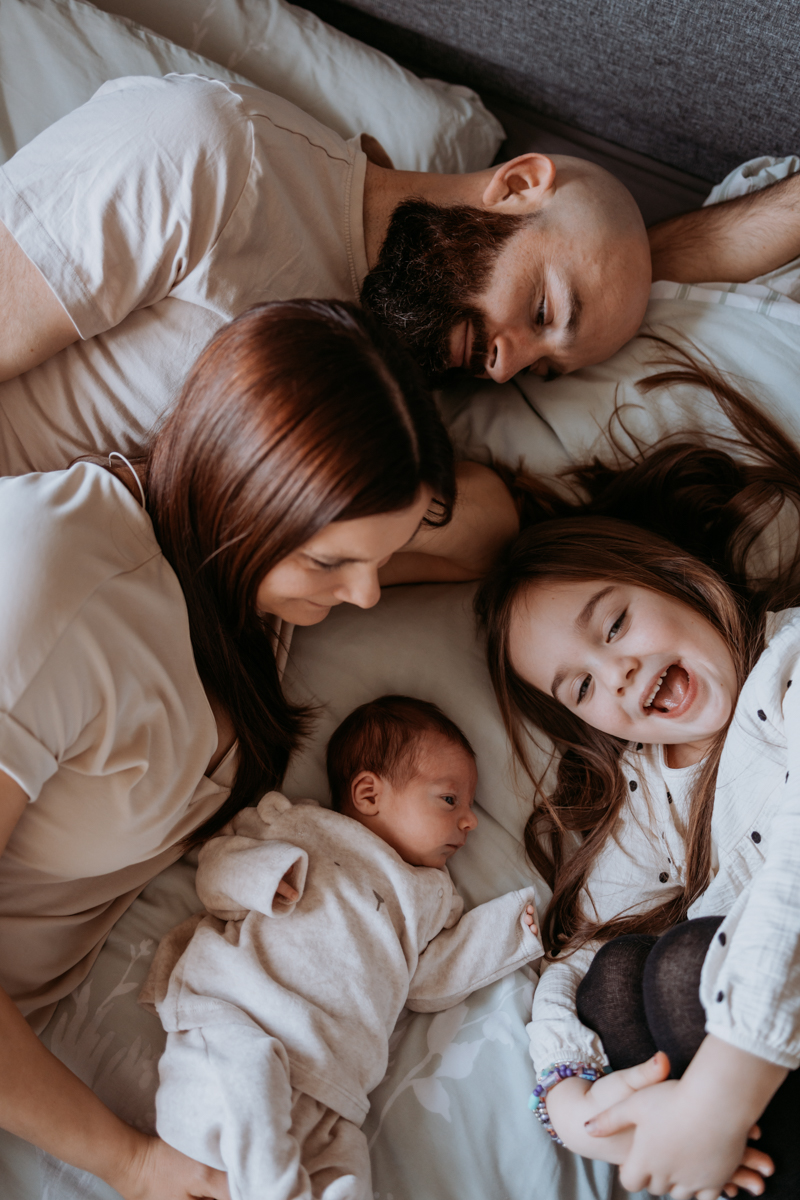 A family lying together on a bed, with a mother and father smiling at their two children, one being a newborn and the other a young girl, who is laughing joyfully.