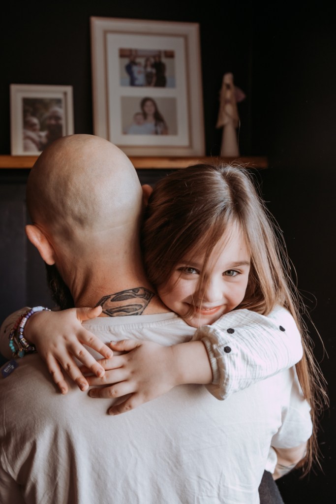 A young girl hugs a bald man from behind, smiling warmly. The background features family photos on a shelf.