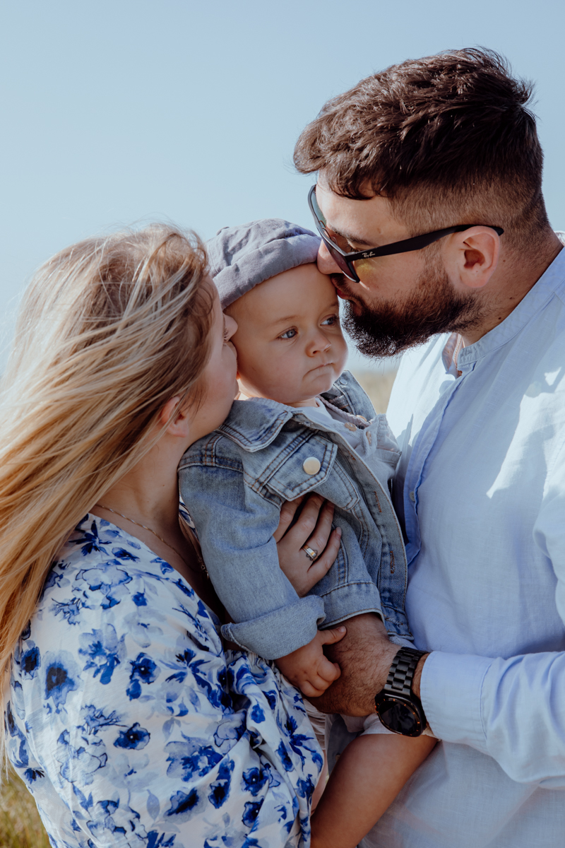 A mother and father kissing their baby on the forehead while the mother holds the baby, who is wearing a denim jacket and a hat. The family is outdoors with a clear blue sky in the background.