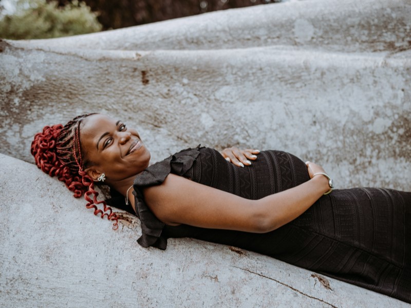 A smiling pregnant woman reclining on a textured surface, wearing a black dress with ruffled sleeves.
