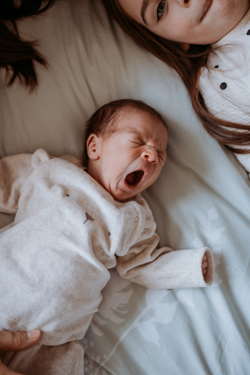 A newborn baby yawning while lying on a bed, wearing a soft, light-colored outfit, with a child's face partially visible in the background.