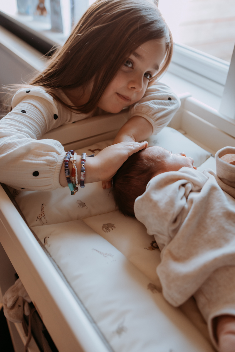 A young girl gently touches the head of a sleeping baby in a crib, with a window in the background. The girl is wearing a cream-colored top with polka dots and colorful bracelets.
