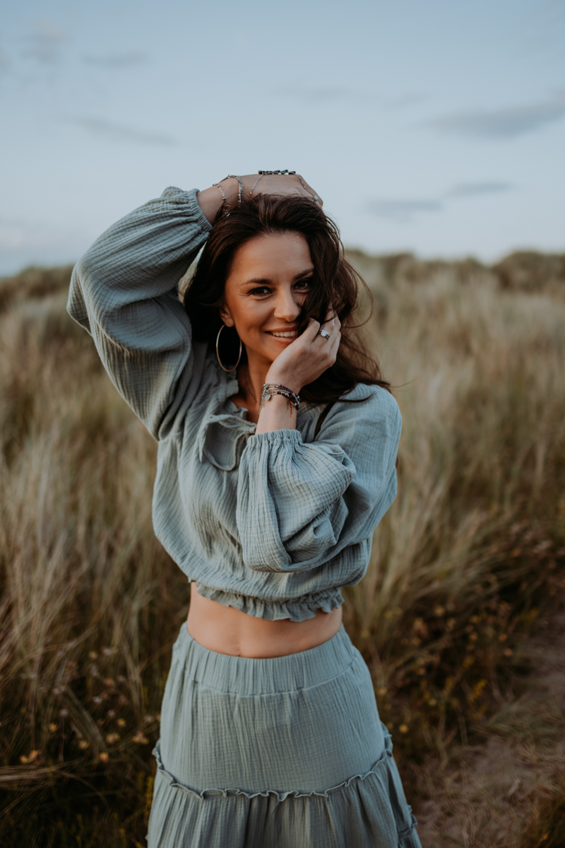 A woman smiling and posing in a meadow, wearing a light green outfit with a cropped top and a flowy skirt, surrounded by tall grass.