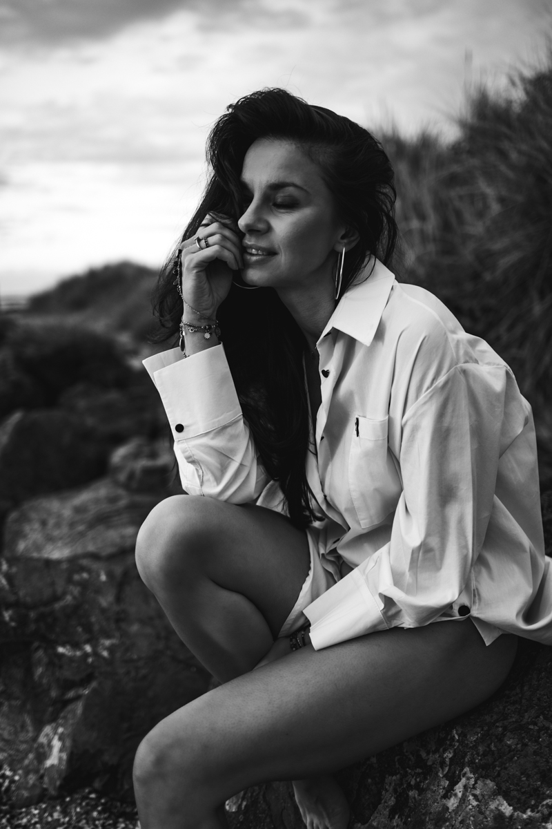A woman seated on rocks, wearing an oversized white shirt, with her hair cascading down. She is smiling softly and has her hand resting on her chin. The image is in black and white, with a serene beach background.