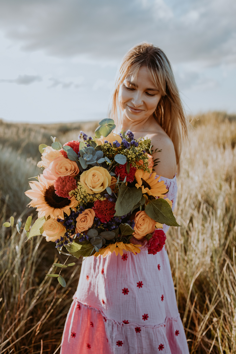 A woman in a floral dress holding a vibrant bouquet of mixed flowers, including sunflowers and roses, standing in a grassy field under a cloudy sky.