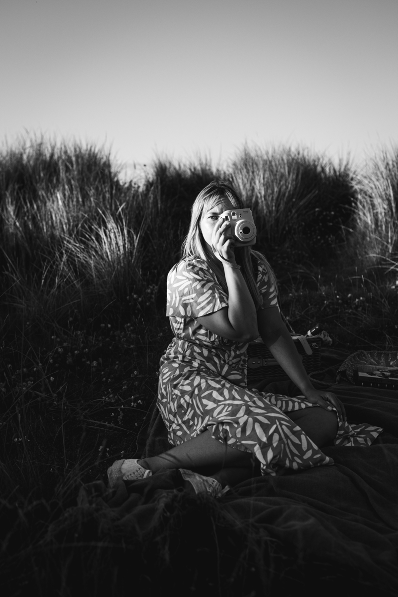 A woman sitting on a blanket in a grassy area, holding a camera and looking directly at the viewer, wearing a patterned dress.