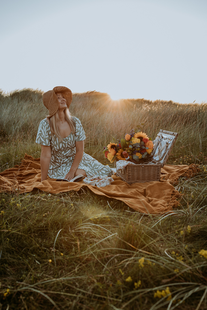 A woman wearing a floral dress and a straw hat sits on a blanket in a grassy field, with a picnic basket and a bouquet of flowers beside her, enjoying a sunset.