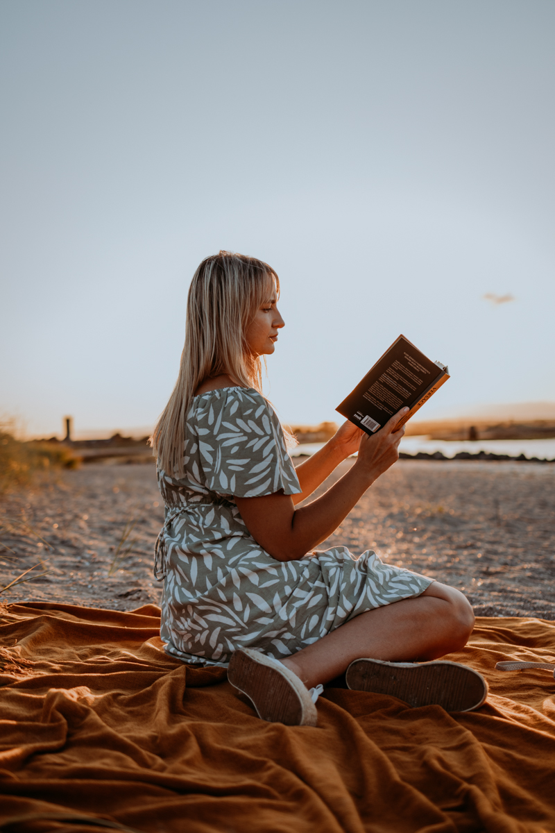 A woman sitting on a sandy beach, reading a book while wearing a light patterned dress. She is perched on an orange blanket, with a serene water view in the background during sunset.