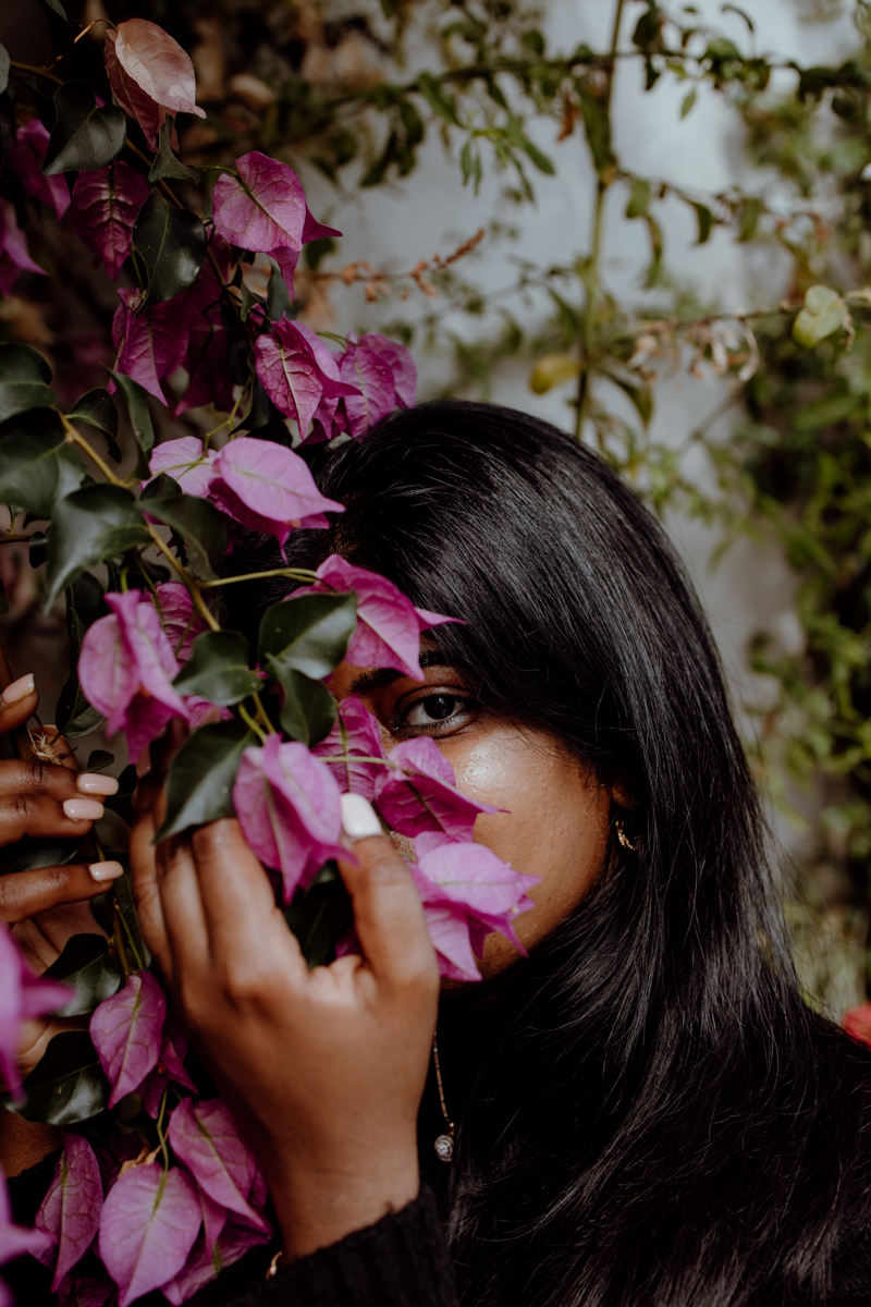 A woman partially hidden behind vibrant purple leaves, showcasing her eye and hand with delicate features, set against a blurred natural background.