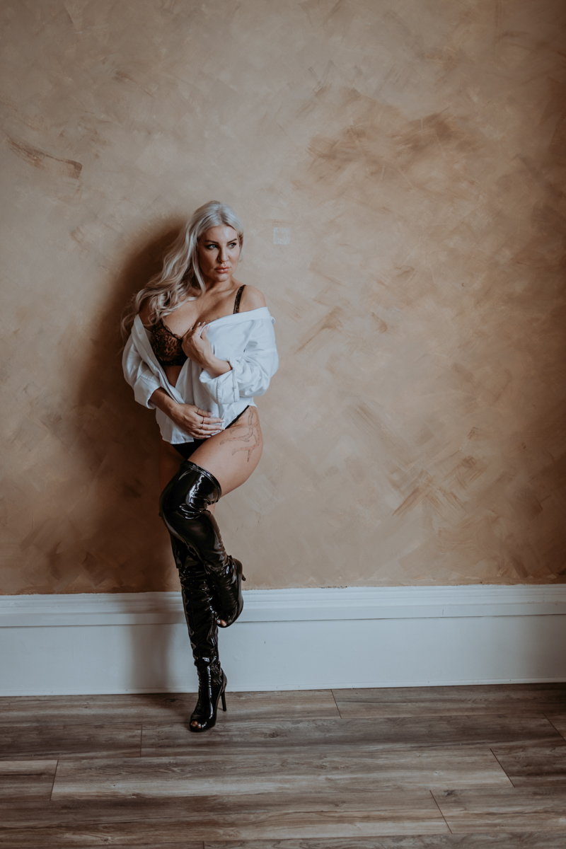 A woman with long, wavy blonde hair poses against a textured beige wall, wearing a black lace bra and high-waisted underwear, paired with a loose white shirt and thigh-high black boots.