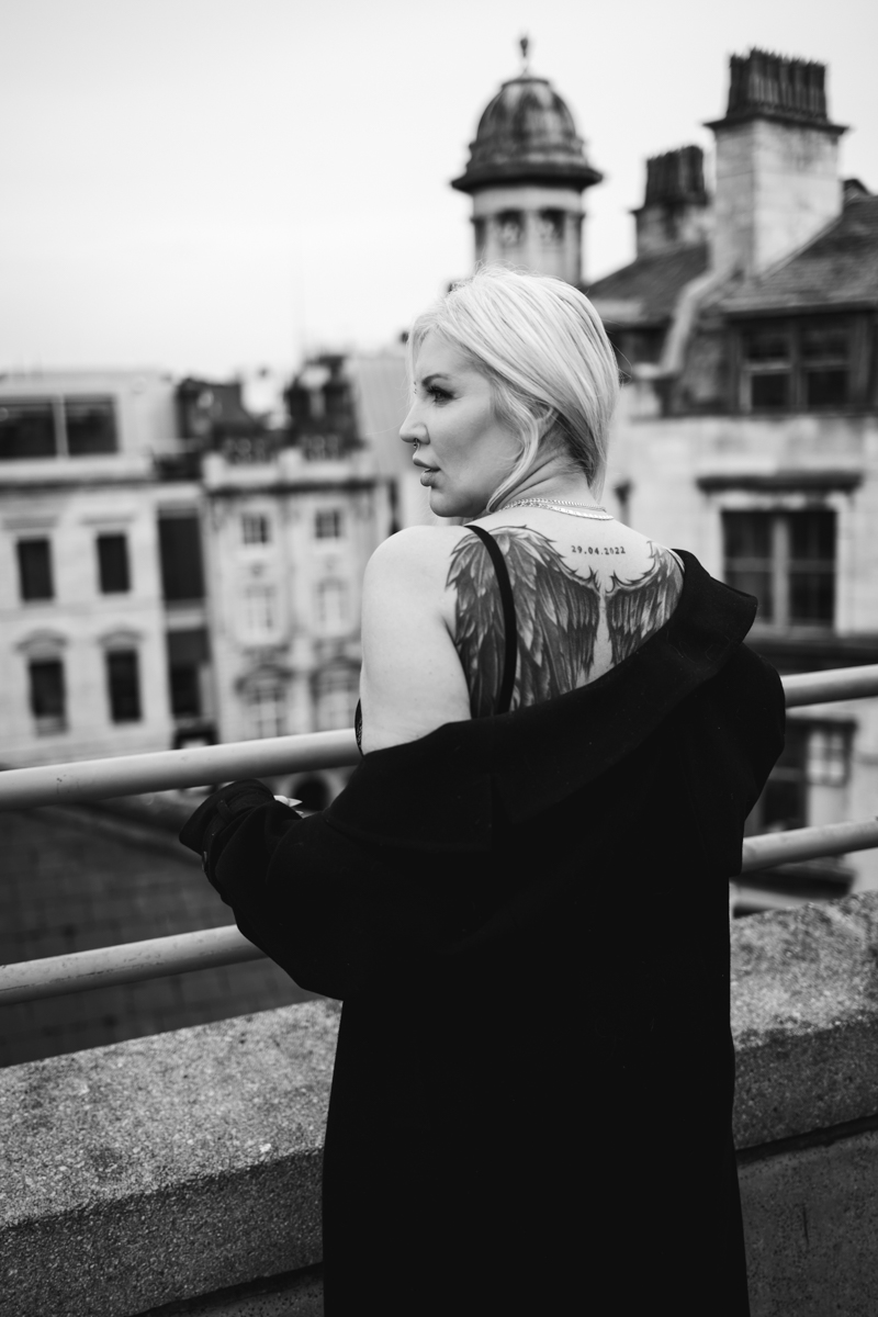 A woman with blonde hair stands on a rooftop, wearing a black coat, looking away from the camera. Her back is partially visible, showcasing a large tattoo of angel wings.