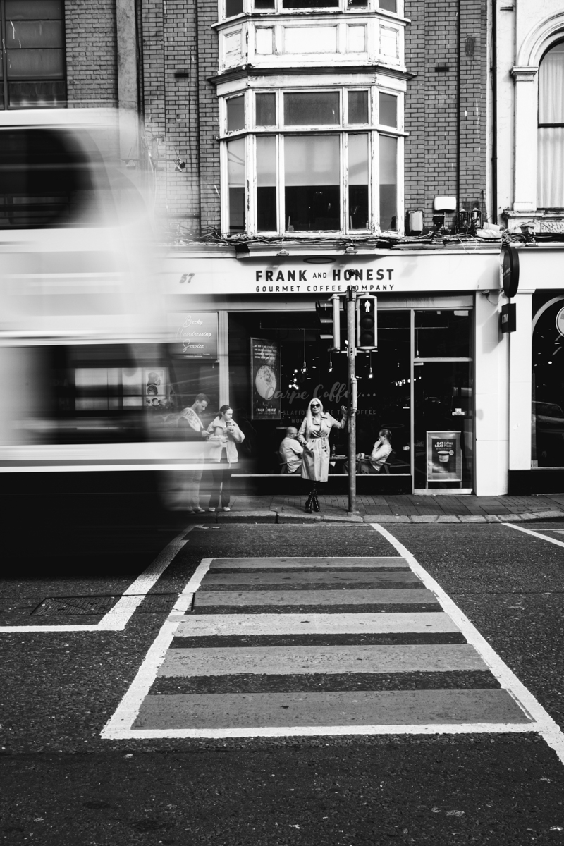 A black and white street scene featuring a coffee shop named 'Frank and Honest'. In the foreground, a pedestrian crossing with people standing on the sidewalk, while a bus passes by in motion.
