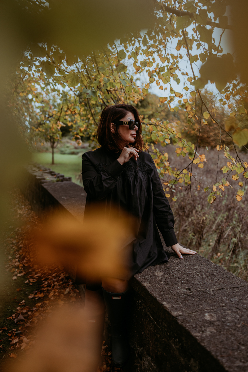 A woman wearing a black dress and sunglasses stands by a stone wall in an autumn setting, surrounded by foliage with yellow leaves.