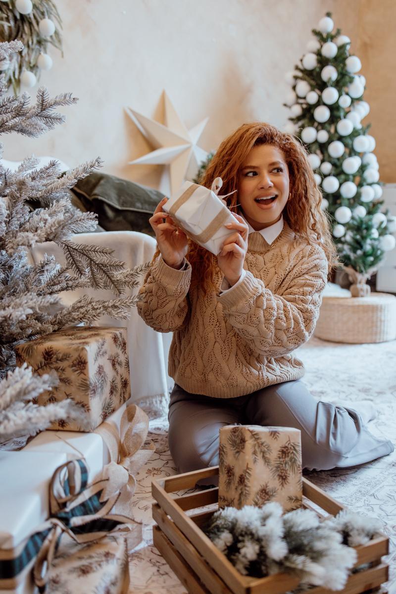 A smiling woman with curly hair wearing a cozy sweater is sitting on the floor surrounded by Christmas gifts and decorations. She holds a small gift box and appears excited amidst a festive setting with a decorated Christmas tree in the background.