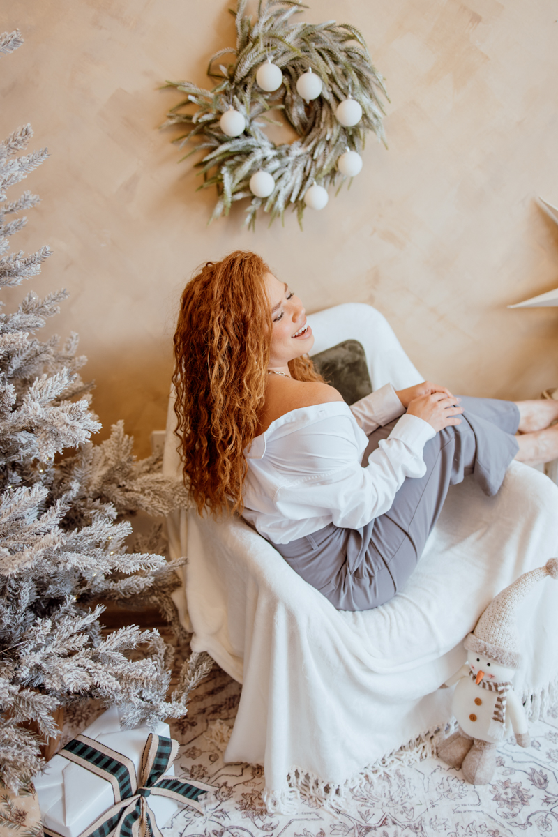 A woman with curly red hair smiles while seated on a white chair, wearing an off-the-shoulder white blouse and gray pants. A snowy Christmas tree and a festive wreath with white ornaments are in the background, along with a wrapped gift and a snowman figurine.