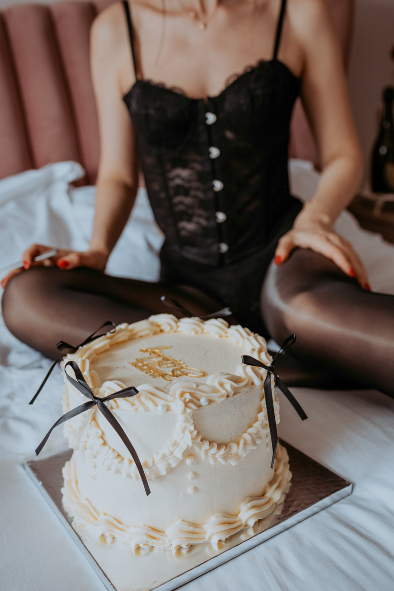 A woman in a black lace bodysuit sitting on a bed with a decorated birthday cake in front of her, featuring cream frosting and black ribbon accents.