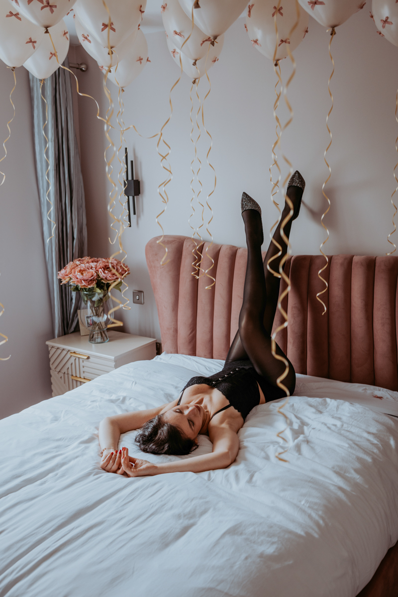 A woman lying on a bed, dressed in black lingerie, with her legs raised in the air. The room is decorated with white balloons with pink bows and festive gold streamers, alongside a bouquet of pink flowers on a nightstand.