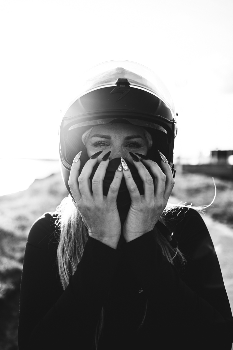 A close-up of a woman wearing a motorcycle helmet, holding a face mask with both hands and showing off long, decorated nails, set against a bright background.