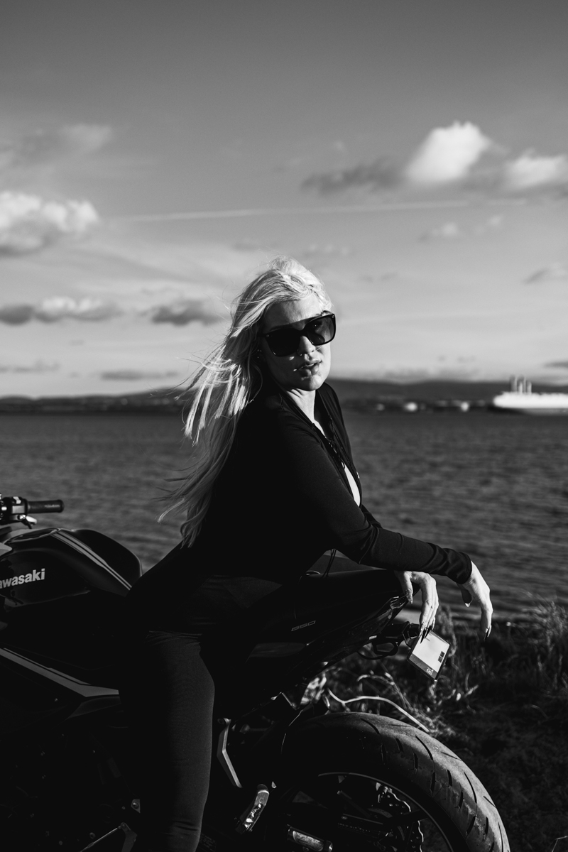 A woman with long blonde hair wearing sunglasses poses on a black motorcycle against a scenic waterfront background, captured in black and white.